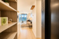 Entry hall of a modern apartment opens to a bright living room with a beige sofa and city skyline through large windows, shelves on the left and an elevator door on the right.