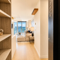 Entry hall of a modern apartment opens to a bright living room with a beige sofa and city skyline through large windows, shelves on the left and an elevator door on the right.