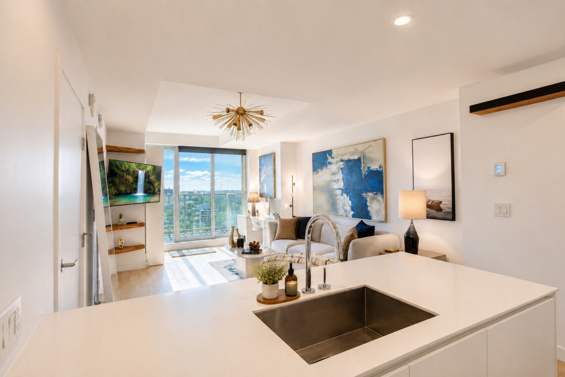 Open-concept living area with a white kitchen island, stainless sink, sofa, abstract blue art, and a balcony view of the city.