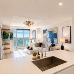 Open-concept living area with a white kitchen island, stainless sink, sofa, abstract blue art, and a balcony view of the city.
