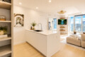 Bright open-plan kitchen and living room with a white island, sink, plants, and a city skyline visible through floor-to-ceiling windows.