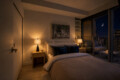 Cozy hotel bedroom at night with a king bed, warm lamps on nightstands, and a balcony showing a city skyline and crescent moon outside.