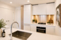 Modern white kitchen with a stainless-steel sink island in the foreground and a built-in oven and gas range along the back wall.