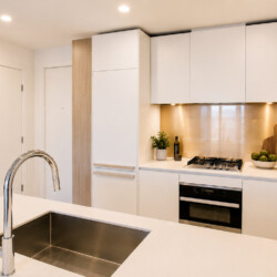 Modern white kitchen with a stainless-steel sink island in the foreground and a built-in oven and gas range along the back wall.