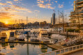 Sunset over a busy marina with anchored yachts and a skyline of modern buildings in the background.