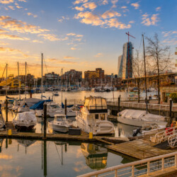 Sunset over a busy marina with anchored yachts and a skyline of modern buildings in the background.
