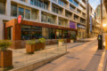 Modern storefronts along a sunny street, including Tim Hortons and London Drugs, with glass windows and planters in front of a tall building complex.