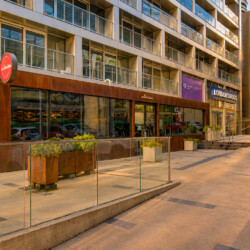 Modern storefronts along a sunny street, including Tim Hortons and London Drugs, with glass windows and planters in front of a tall building complex.