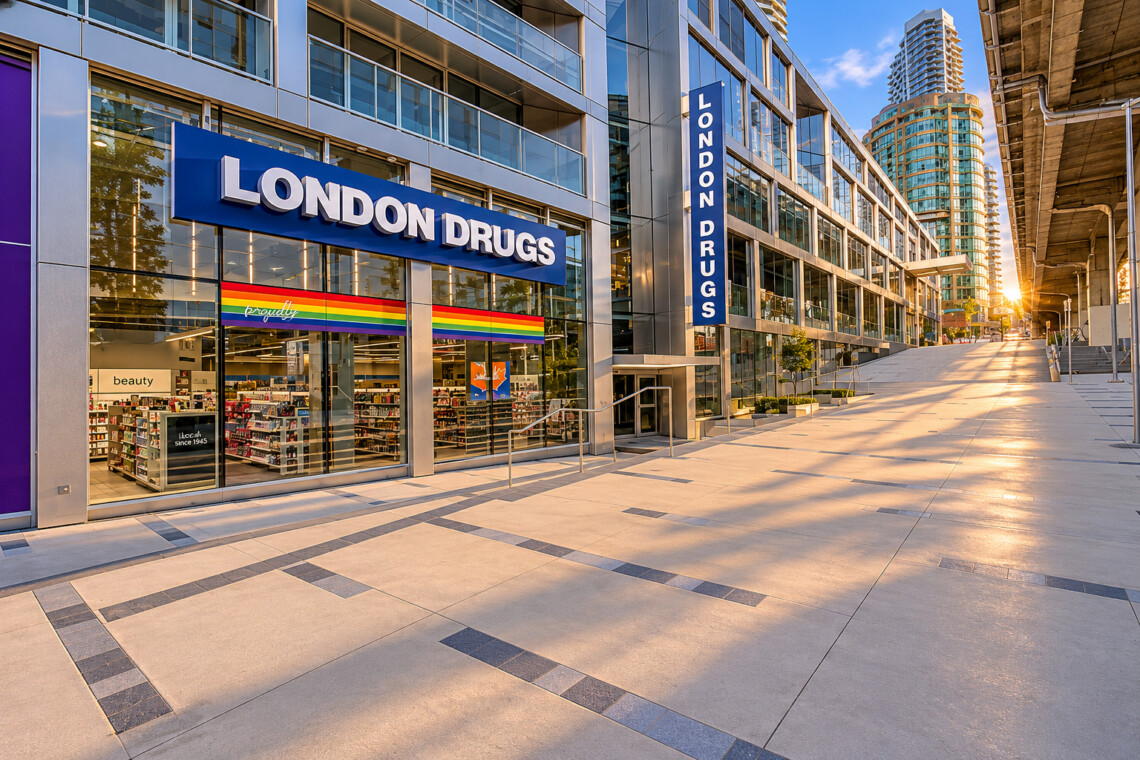 London Drugs storefront in a modern glass building complex with a rainbow Pride flag in the window; sunlit walkway and elevated railings.