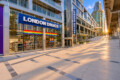 London Drugs storefront in a modern glass building complex with a rainbow Pride flag in the window; sunlit walkway and elevated railings.