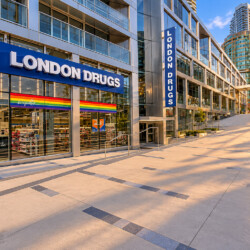 London Drugs storefront in a modern glass building complex with a rainbow Pride flag in the window; sunlit walkway and elevated railings.
