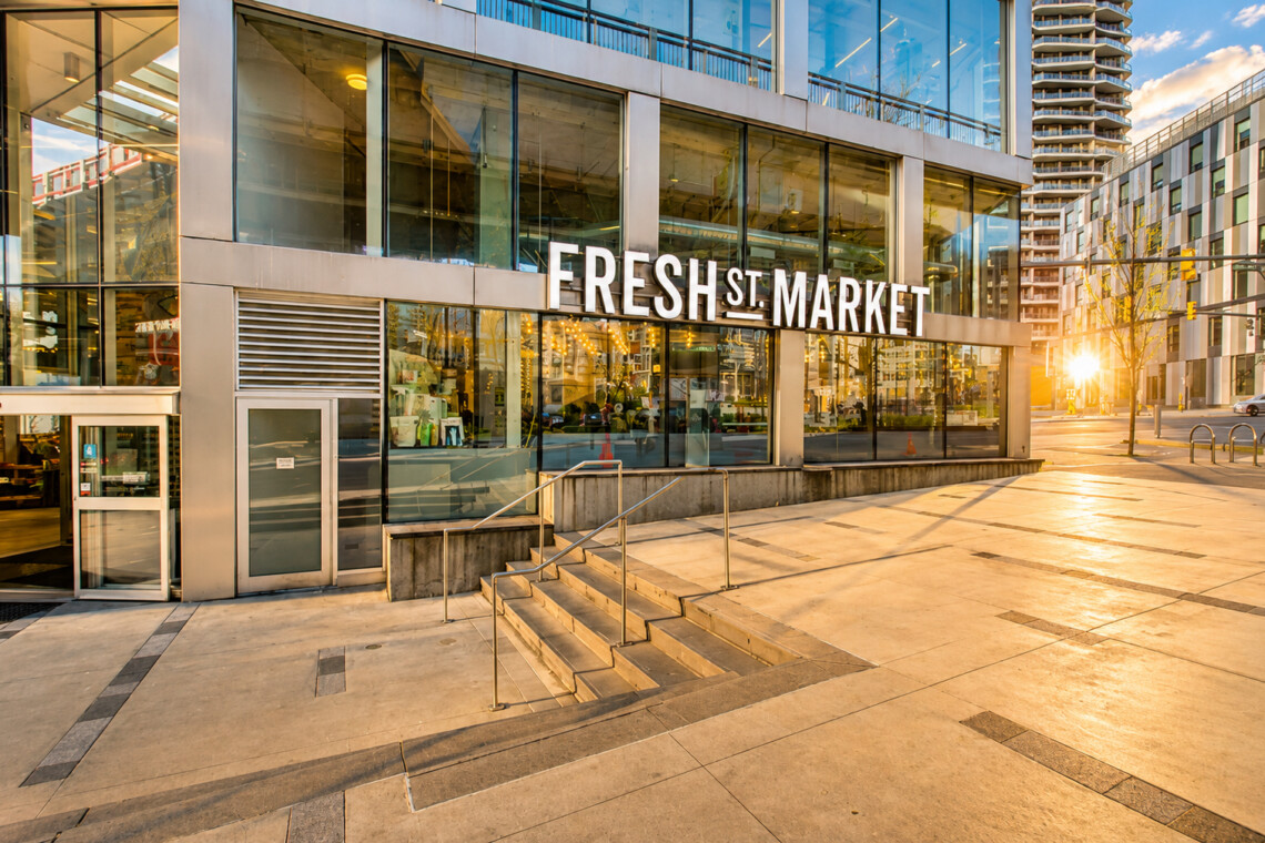 Modern glass-front Fresh Market storefront in a sunny urban plaza with stairs and metal railings outside