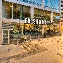 Modern glass-front Fresh Market storefront in a sunny urban plaza with stairs and metal railings outside