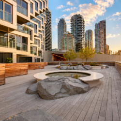 Rooftop courtyard with circular planter water feature, wooden deck, and modern high-rise buildings at sunset.