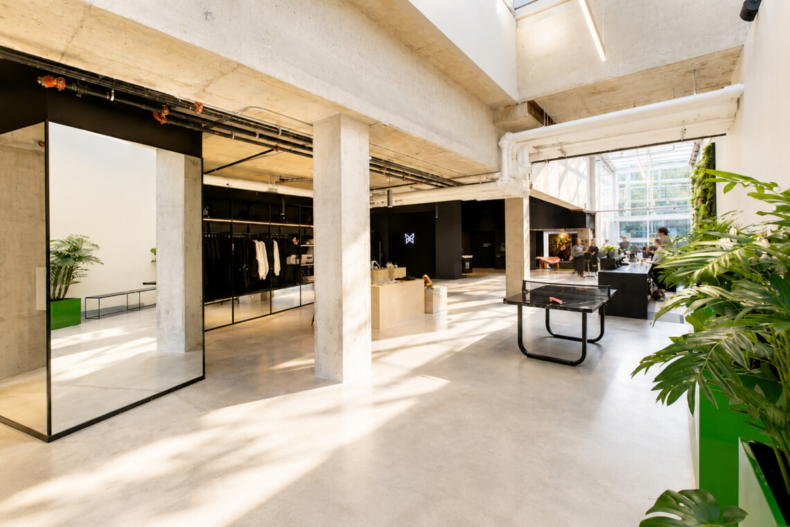 Open-plan retail space with concrete pillars, clothing racks along the left, a ping-pong table in the foreground, and large windows flooding the area with light.