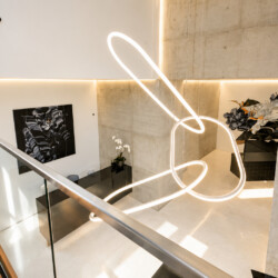 Modern interior lobby with a large glowing abstract neon sculpture hanging above a glass railing and polished floor.