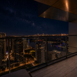 Night skyline viewed from a balcony: lit high-rise buildings, a harbor, and a starry sky.