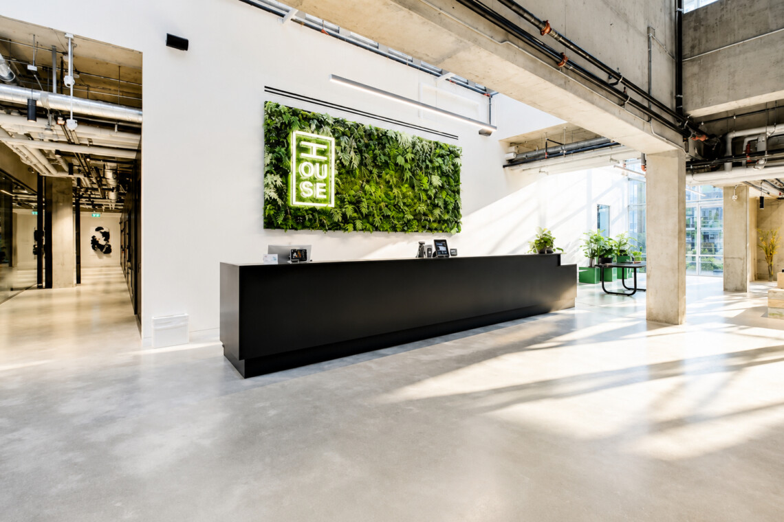 Modern office reception: long black counter under a green plant wall with a glowing logo, sunlit open space.