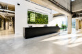 Modern office reception: long black counter under a green plant wall with a glowing logo, sunlit open space.