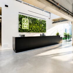Modern office reception: long black counter under a green plant wall with a glowing logo, sunlit open space.