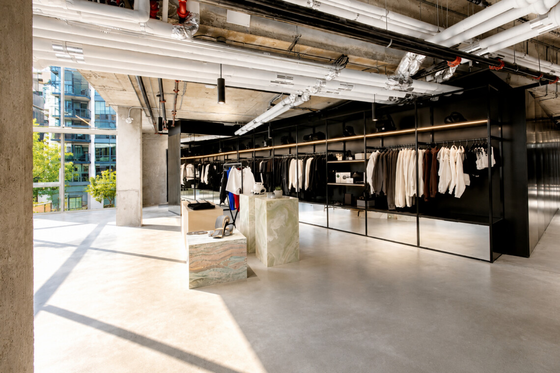 Open-plan clothing store with black metal racks full of white and neutral coats, concrete columns, and large sunlit windows.