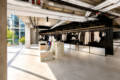 Open-plan clothing store with black metal racks full of white and neutral coats, concrete columns, and large sunlit windows.