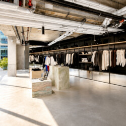Open-plan clothing store with black metal racks full of white and neutral coats, concrete columns, and large sunlit windows.