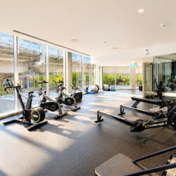 Bright modern gym with rowers and stationary bikes by large windows overlooking an urban exterior likely a transportation area outside the glass walls.