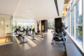 Bright gym with rowers and stationary bikes along glass walls, city buildings visible outside the windows.
