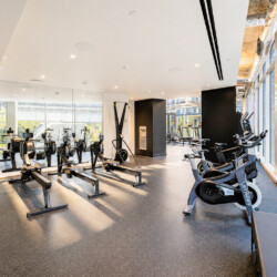 Bright gym with rowers and stationary bikes along glass walls, city buildings visible outside the windows.