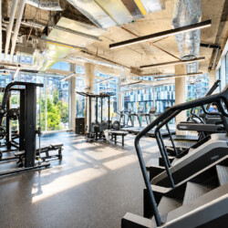 Modern gym interior with treadmills in the foreground, weight machines, exposed ducts, and large windows with a city view.
