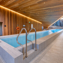 Indoor pool in a modern wooden atrium with warm ceiling lights and stainless handrails beside a calm blue pool