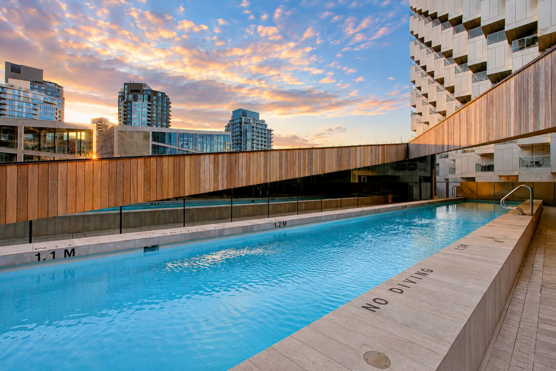 Rooftop swimming pool with wooden railing and modern buildings against a sunset sky in the background