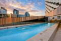 Rooftop swimming pool with wooden railing and modern buildings against a sunset sky in the background