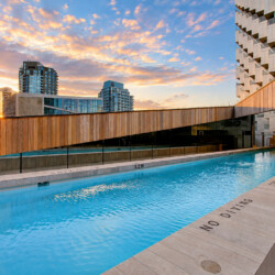 Rooftop swimming pool with wooden railing and modern buildings against a sunset sky in the background