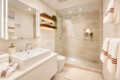 Modern bathroom with a double-sink vanity, backlit mirror, and a glass-enclosed shower with a rainfall showerhead; beige tiles and white towels with brown stripes.