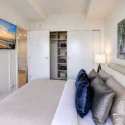 Modern bedroom with a wall-mounted flat-screen TV, open closet shelves, and a beige bed with multiple pillows.
