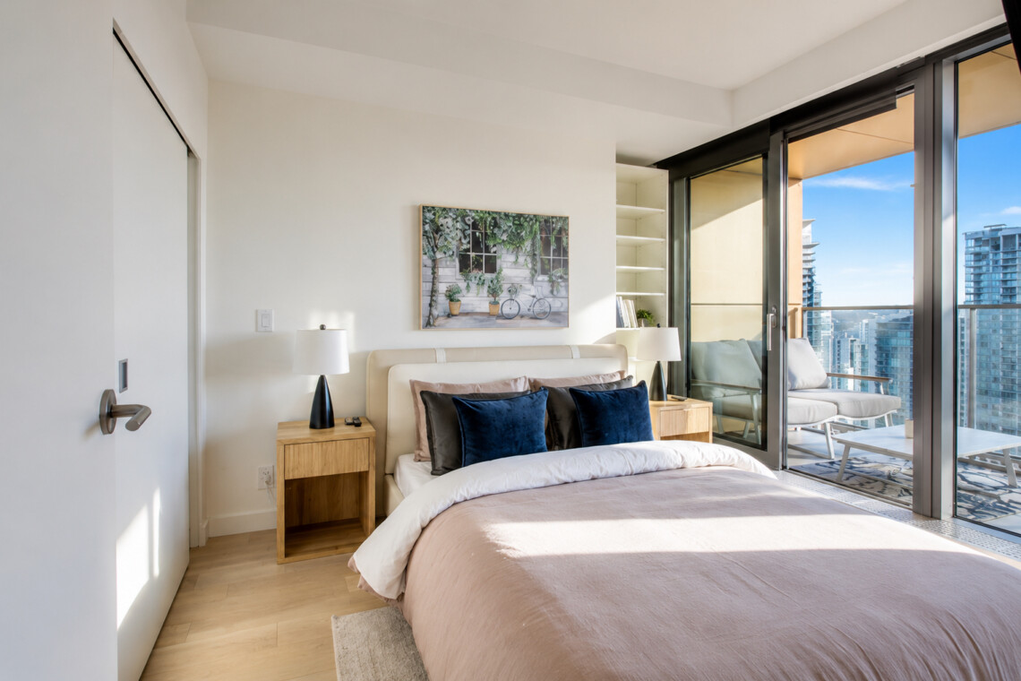 Bright bedroom with a pink duvet, blue and black pillows, two wooden nightstands and lamps, and a balcony with city view.
