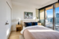 Bright bedroom with a pink duvet, blue and black pillows, two wooden nightstands and lamps, and a balcony with city view.