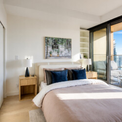 Bright bedroom with a pink duvet, blue and black pillows, two wooden nightstands and lamps, and a balcony with city view.