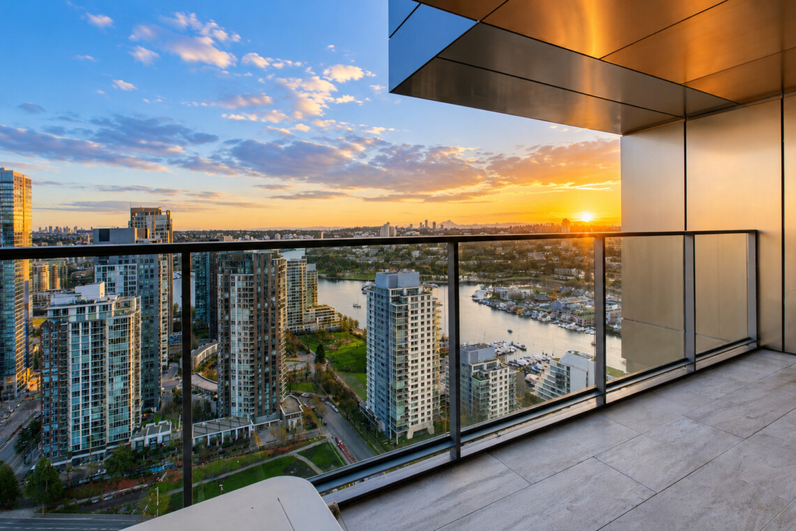 Sunset over a city skyline viewed from a high balcony with a glass railing and river below.