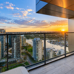 Sunset over a city skyline viewed from a high balcony with a glass railing and river below.