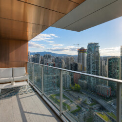 Balcony lounge with white cushioned seating and a glass railing overlooking a dense city skyline under a sunny sky.