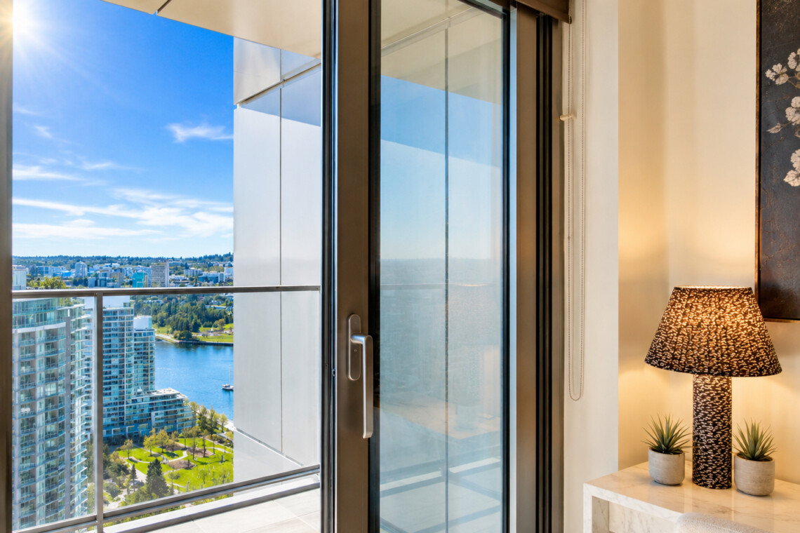 Hotel room with a sliding glass door open to a balcony, offering a river and city skyline view on a sunny day.