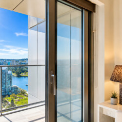 Hotel room with a sliding glass door open to a balcony, offering a river and city skyline view on a sunny day.