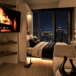 Modern bedroom at night with a lit fireplace displayed on a wall-mounted TV and a bed by a window overlooking a city skyline.