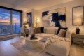 Cozy modern living room with a beige sectional, abstract blue painting, and a sunset city view through large windows.