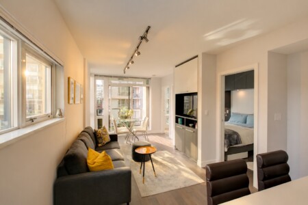 Open-plan living room with a gray sofa, yellow cushion, and a small orange-trim table, bathed in natural light from large windows and a balcony door.