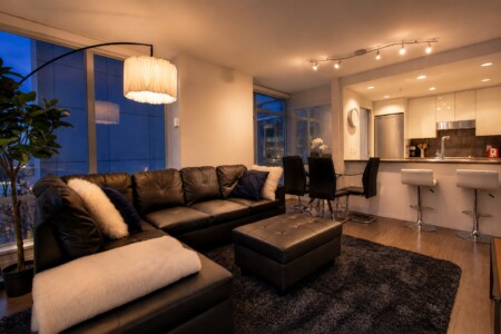 Open-concept living area with a black leather sectional, tufted ottoman, white throws, and a modern kitchen with a breakfast bar and stools.