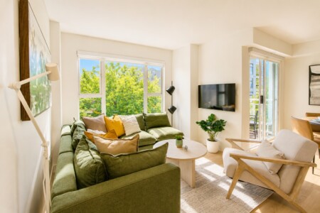 Bright living room with an olive-green sectional, colorful throw pillows, and a round wooden coffee table by a large window overlookings trees, plus a wall-mounted TV nearby.